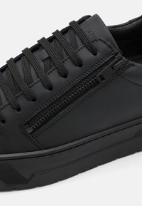 Baskets noires avec lacets ronds noirs et fermeture éclair latérale sur une tige en cuir synthétique lisse et une semelle en caoutchouc épaisse.