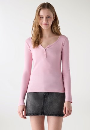 Młoda kobieta o długich włosach, ubrana w jasnoróżową bluzkę z długim rękawem i guzikami oraz czarną dżinsową mini spódniczkę, stoi na jednolitym tle.