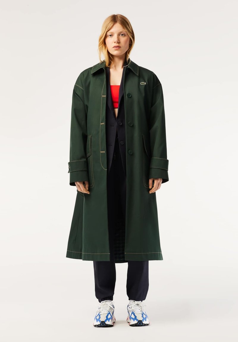 Femme portant un long manteau vert foncé sur un costume noir et un crop top rouge, associé à un pantalon noir et des baskets blanches avec des détails bleus.