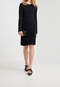 Femme portant une robe longue noire à manches longues, des escarpins noirs à bouts pointus avec des nœuds à la cheville, tenant une petite pochette noire contre un fond blanc uni.