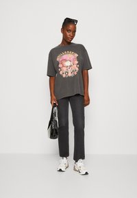Camiseta gris de gran tamaño con un gráfico colorido de calavera y flores, combinada con jeans negros de pierna recta y zapatillas multicolores. Bolsa negra.