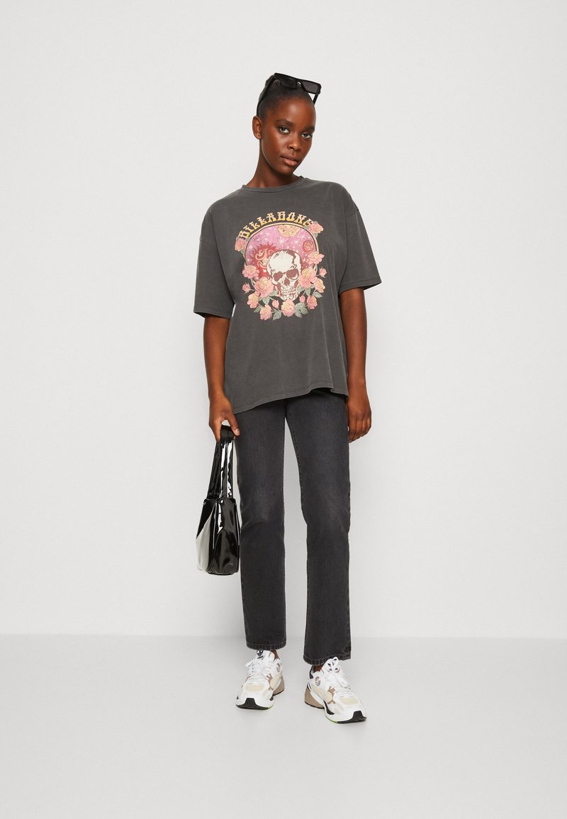 T-shirt oversize gris avec un graphique de crâne coloré et floral, associé à un jean noir à jambe droite et des baskets multicolores. Sac à main noir.