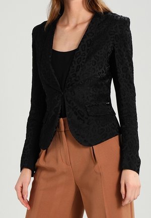 Kvinde iført en tætsiddende sort leopardmønstret blazer over en sort top med højtailede, viddevævede brune bukser.