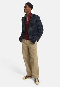 Marine gestreifter Blazer, burgunderfarbener Reißverschluss-Pullover, beiges Hosen und dunkelbraune Sneakers mit cremefarbenen Akzenten. Schmale Passform, geschichteter Look.