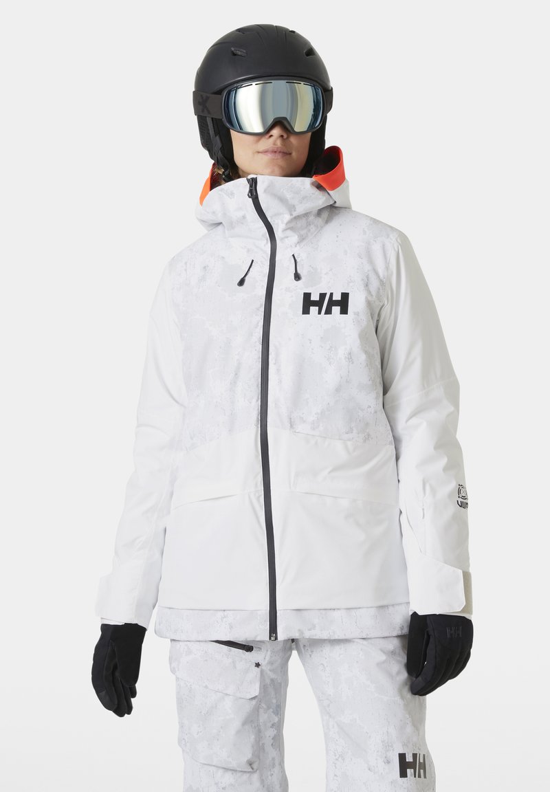 Hvit skijakke med strukturert, mønstret overdel; har svarte detaljer, glidelås foran og avtagbar hette. Svarte hansker.