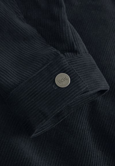 Černá kordurová látka se žebrovanou strukturou a knoflíkovým detailem s nápisem "WM." na manžetě. Měkký, odolný materiál, dobře padnoucí design.