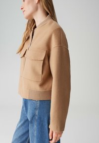 Giacca corta color beige in misto lana con cerniera, dotata di due tasche frontali e polsini e orlo a costine, abbinata a jeans di denim blu.