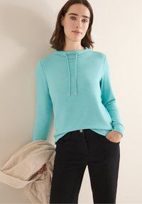 Mujer con una blusa de manga larga azul claro y pantalones negros, sosteniendo una chaqueta de forro polar beige, de pie contra una pared lisa.