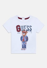 Hvid T-shirt med korte ærmer, der har et grafisk motiv af en bjørn iført en blå jakke og jeans, med teksten "GUESS CALIFORNIA" trykt på forsiden.