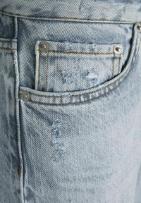 Šviesiai mėlyni džinsai su pažeista tekstūra, su suplėšytomis detalėmis, sidabriniais akcentais ir kontrastine siūle šalia užpakalinės kišenės.