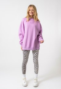 Nagy lila pulóver laza szabással és hímzett felirattal, formás leopárdmintás leggingsszel és fehér vastag talpú sportcipőkkel párosítva.
