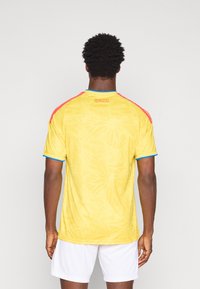 Κίτρινη αθλητική μπλούζα με σχέδιο πεταλούδας, κόκκινες και μπλε λεπτομέρειες στους ώμους και κείμενο "ΚΟΛΟΜΒΙΑ" κάτω από το λαιμό στο πίσω μέρος. Κοντά μανίκια.