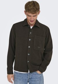 Chaqueta marrón abotonada con cuello, dos bolsillos frontales, costuras de acento azules y botones transparentes. Lleva puesta sobre una camiseta blanca.
