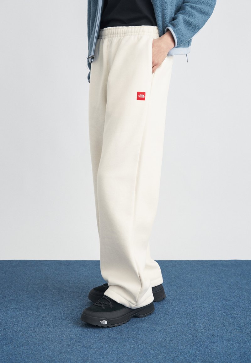 Άτομο που φοράει κρεμ φόρμες με κόκκινο λογότυπο, μαύρα παπούτσια, μπλε μπουφάν, στέκεται σε μπλε χαλί.