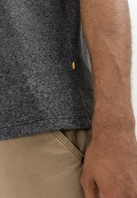 Grijze getextureerde t-shirt met een klein geel label aan de linkerkant, gecombineerd met beige broeken. Focus op de stof en het kleurcontrast.