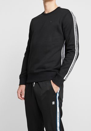 Czarna bluza z okrągłym dekoltem, z białymi paskami po bokach i logo na piersi. Wykonana z gładkiej tkaniny z prążkowanymi mankietami i dołem.