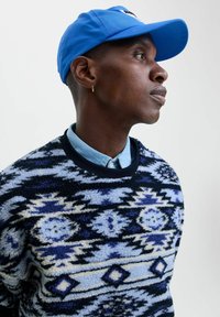 Gebreide trui in blauw en zwart geometrisch patroon met crèmekleurige accenten, gedragen over een lichtblauwe overhemd met kraag. Blauwe pet op het hoofd.