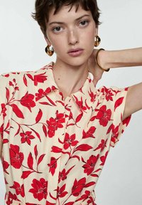 Jonge vrouw met kort haar, gekleed in een crèmekleurig shirt met rode bloemenprint, grote gouden oorbellen en een gouden armband, poserend met haar hand achter haar hoofd.