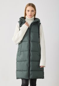 Ermeløs, mørkegrønn puffer vest med glidelås foran, hette og sidelommer. Har en glatt tekstur og sydd paneldesign.