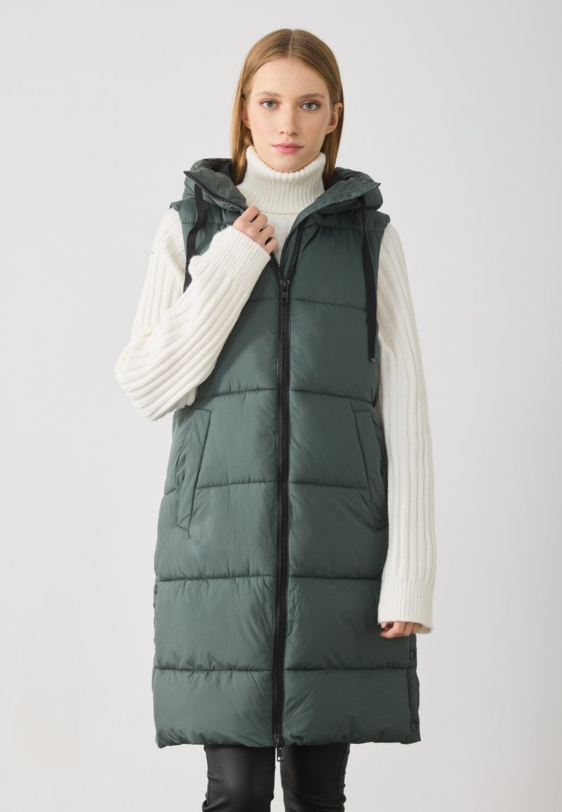 Ermeløs, mørkegrønn puffer vest med glidelås foran, hette og sidelommer. Har en glatt tekstur og sydd paneldesign.