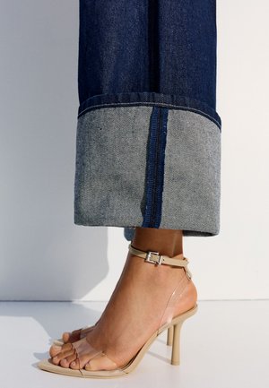 Enkel met donkerblauwe omgeslagen spijkerbroek en beige hoge hak sandalen met doorzichtige bandjes en een enkelgesp.