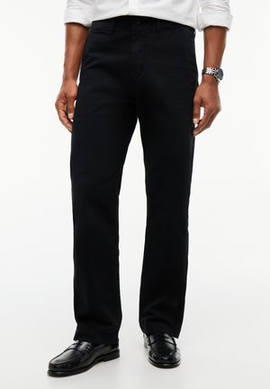 Mann trägt schwarze Hose mit geradem Bein, schwarze Lederschuhe, weißes Hemd mit hochgekrempelten Ärmeln und eine silberne Armbanduhr.