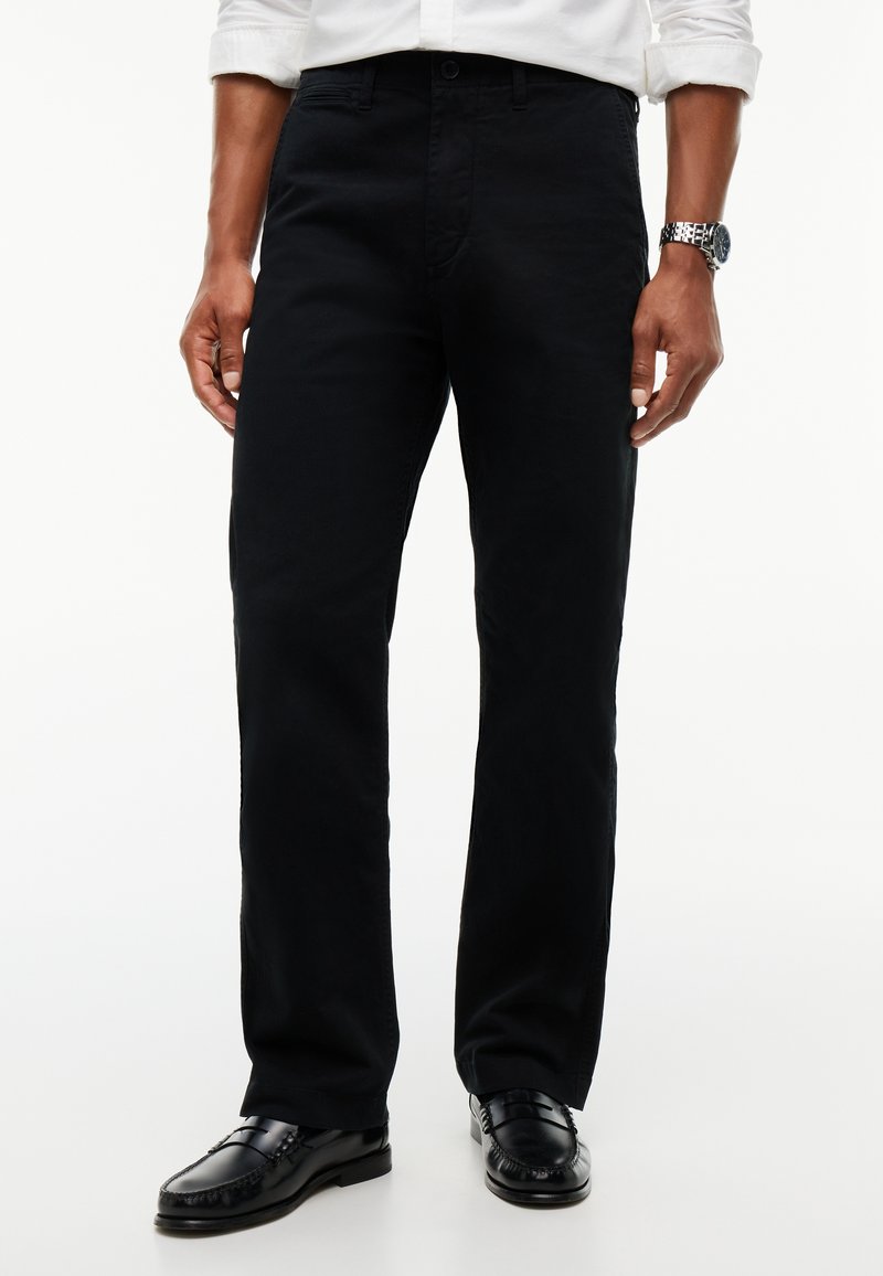 Homme portant un pantalon noir à jambes droites, des chaussures en cuir noires, une chemise blanche aux manches retroussées et une montre-bracelet argentée.