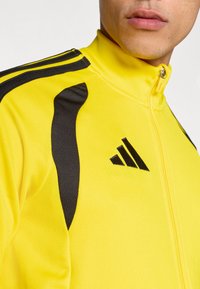 Κίτρινο αθλητικό μπουφάν με φερμουάρ, με μαύρες λεπτομέρειες και υφή υφάσματος. Εμφανές λογότυπο Adidas στο στήθος, με μαύρες ρίγες στους ώμους.