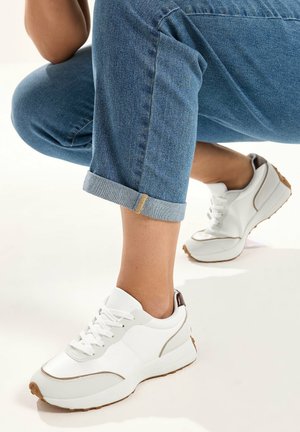 Persona con jeans azules remangados y zapatillas blancas con detalles marrones, en posición agachada sobre un fondo blanco.