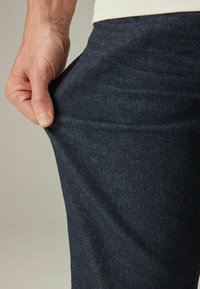 Donkerblauwe denim broek met een gladde textuur, vastgehouden bij de tailleband om de rek en pasvorm te tonen. Versterkte stiksels aan de randen.