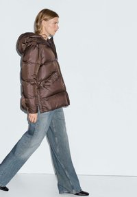 Donna che cammina verso sinistra indossando un piumino marrone con cappuccio, jeans blu a gamba larga e scarpe nere a punta su uno sfondo chiaro e uniforme.