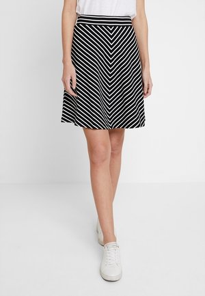 Kvinde iført en sort-hvid striped A-formet nederdel med en hvid top og hvide sneakers, stående mod en enkelt lys baggrund.