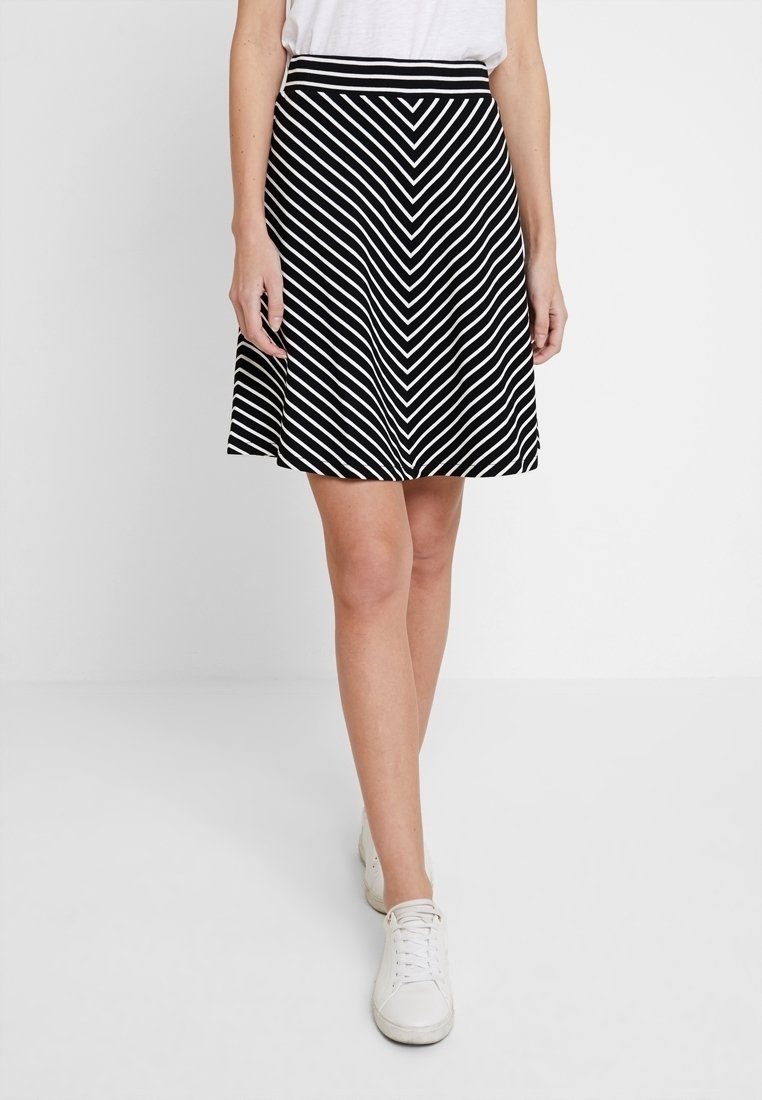 Femme portant une jupe trapèze rayée noire et blanche avec un top blanc et des baskets blanches, debout devant un fond clair uni.
