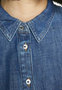 Lähikuva henkilöstä, joka pukeutunut siniseen farkkupaitaan, jossa on napit kauluksessa ja näkyvissä ruskeat ompelusaumat.