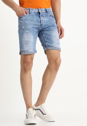 Lichtblauwe denim shorts met opgerolde boorden, voorzien van verweerde details en vijf zakken. Gecombineerd met een effen oranje T-shirt en witte sneakers.