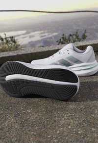 Chaussures de course blanches avec semelle crantée noire posées sur une surface pavée extérieure près d’un rebord en béton avec un paysage flou en arrière-plan.