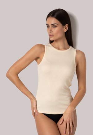 Donna con capelli castani lisci che indossa un top beige senza maniche a costine e biancheria nera, in posa davanti a uno sfondo chiaro uniforme.