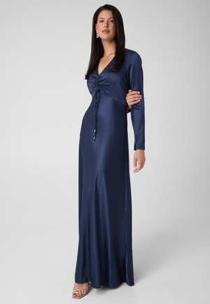 Femme aux longs cheveux foncés portant une robe longue en satin bleu marine à manches longues avec un détail froncé à l'avant et des talons à brides sur un fond uni.