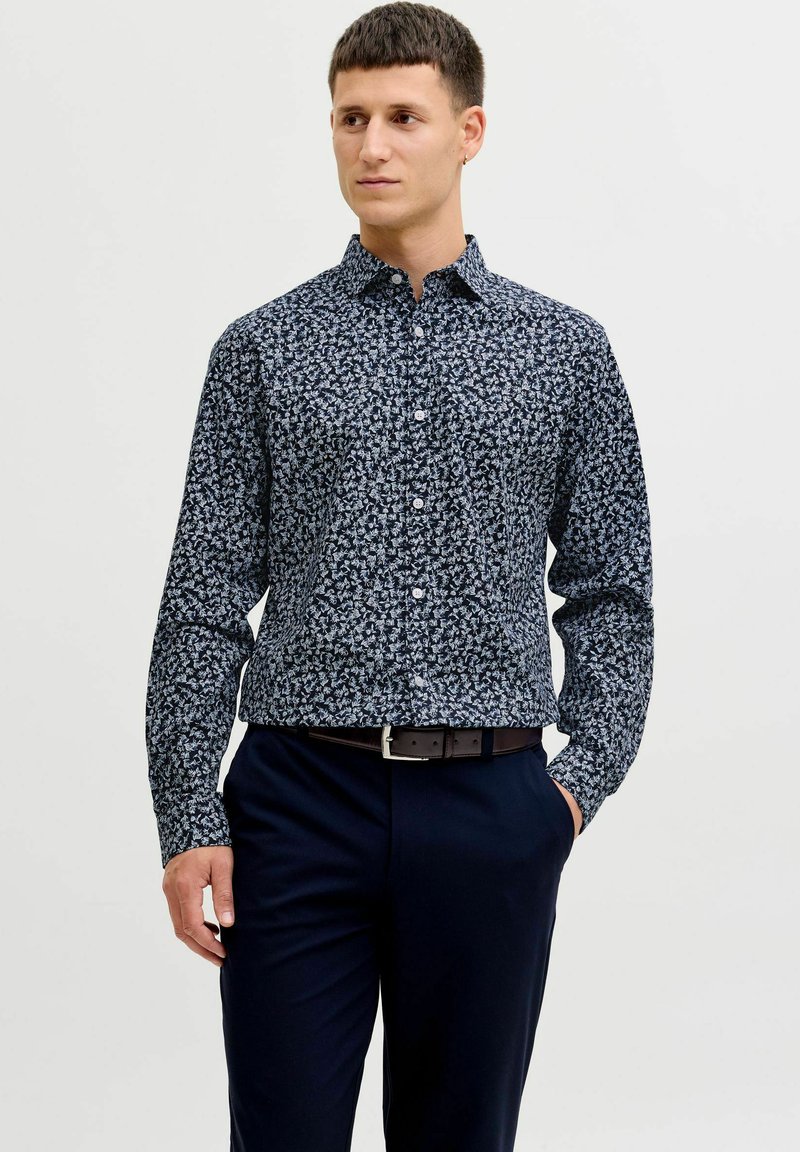 Mees, kes kannab tumelasinist triiksärki helelillelise mustriga, mis on sisse lükatud tumesinineisse pükstesse, millel on must vöö, seisab ühevärvilise tausta ees.