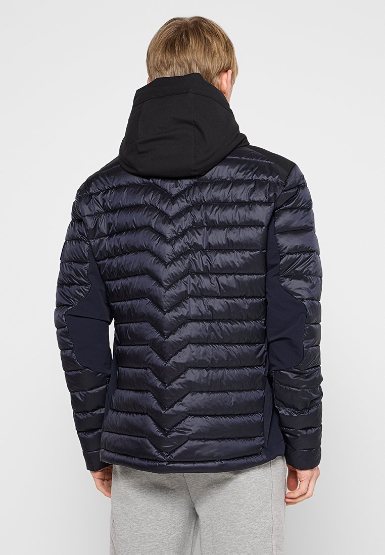 Marineblaue Pufferjacke mit Chevron-Muster, ausgestattet mit einer schwarzen Kapuze und dehnbaren schwarzen Seitenteilen. Glatte Textur und isoliertes Design.