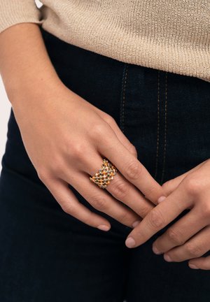 Manos entrelazadas a la altura de la cintura, con vaqueros oscuros y suéter beige, llevando un anillo de malla dorada con cuentas de colores en el dedo medio.