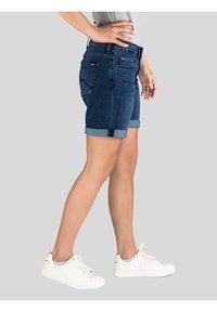 Donkerdenim shorts met opgerolde manchetten, voorzien van achterzakken en een getailleerd ontwerp. G gedragen met witte sneakers tegen een neutrale achtergrond.