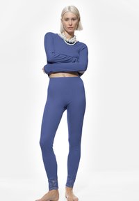 Femme aux cheveux blonds courts, portant un crop top bleu à manches longues et des leggings assortis à des poignets en dentelle, accessoirisée de colliers en perles superposés.