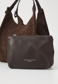 Bruine suède schoudertas met twee handvatten, gecombineerd met een kleine donkerbruine leren etui met een zilveren rits en logo.