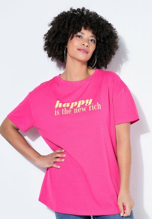 Frau mit lockigem Haar und Creolen, die ein pinkfarbenes T-Shirt mit gelbem Text "Glücklich ist das neue Reich" trägt und mit einer Hand in der Hüfte posiert.