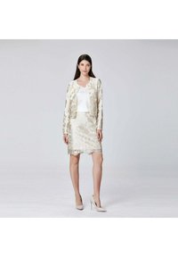 Femme portant une jupe en jacquard floral ivoire et une veste assortie avec un haut blanc et des talons hauts beiges, se tenant devant un fond uni.