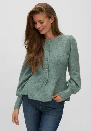 Jeune femme aux longs cheveux bruns portant un pull vert boutonné et un jean bleu, souriante et regardant vers le bas avec une main dans la poche.