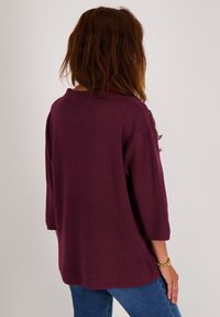 Vrouw met lang bruin haar, draag een losse, driekwart mouwen bordeauxrode top en blauwe jeans, staand tegen een effen lichte achtergrond.