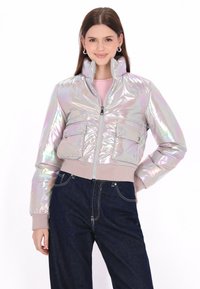 Irydescentna kurtka bombowiec z wysokim kołnierzem, zamkiem błyskawicznym z przodu, dwoma kieszeniami na piersi oraz ściągaczami przy mankietach, połączona z ciemnoniebieskimi dżinsami.