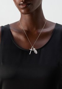 Sølv halskjede med et kors- og fjærvedheng, båret over et svart ermeløst plagg. Glatt tekstur, minimalistisk design og lagdelte kjeder.